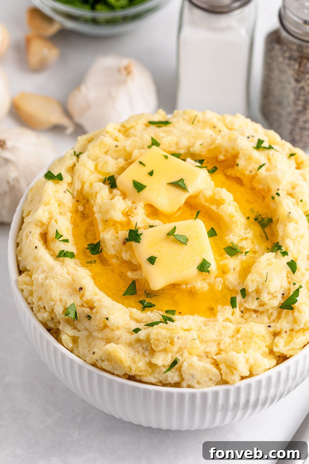 Effortless Creamy Garlic Mashed Potatoes 19 White table with salt and pepper shakers, a handful of garlic cloves, and a white bowl full of Garlic Mashed Potatoes