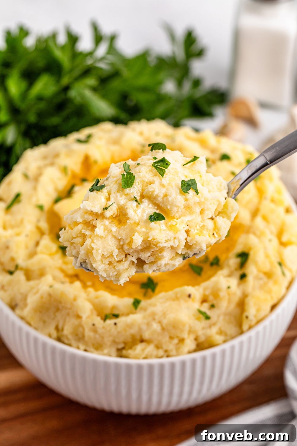 Effortless Creamy Garlic Mashed Potatoes 20 Garlic Mashed Potatoes with a spoon scooping up some out of the white bowl sitting on a wooden cutting board on the table