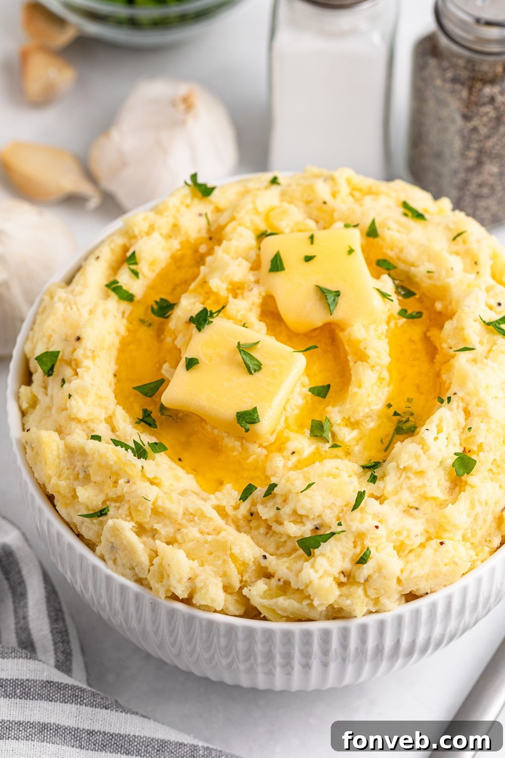 Effortless Creamy Garlic Mashed Potatoes 21 Garlic Mashed Potatoes in a bowl on white table with garlic and a striped towel to side of the bowl