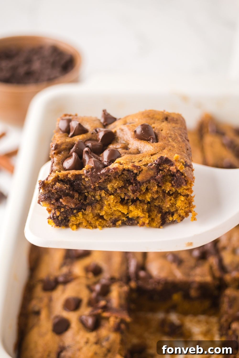 white spatula with a slice of Brown Butter Pumpkin Chocolate Chip Bar on it, and a pan full of bars sitting on table 