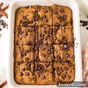 Brown Butter Pumpkin Chocolate Chip Bars in a ceramic casserole dish on table with cinnamon sticks and chocolate chips around table