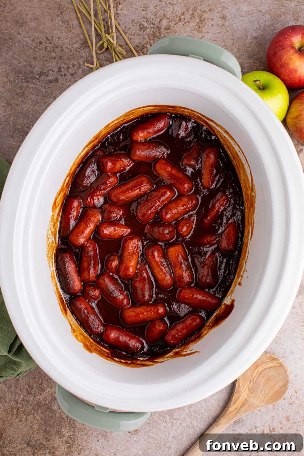 Sweet and Tangy Crockpot Apple Butter Smokies 18 looking down on table with a white Crockpot full of Apple Butter BBQ Little Smokies with apples to the side of table and wooden spoon