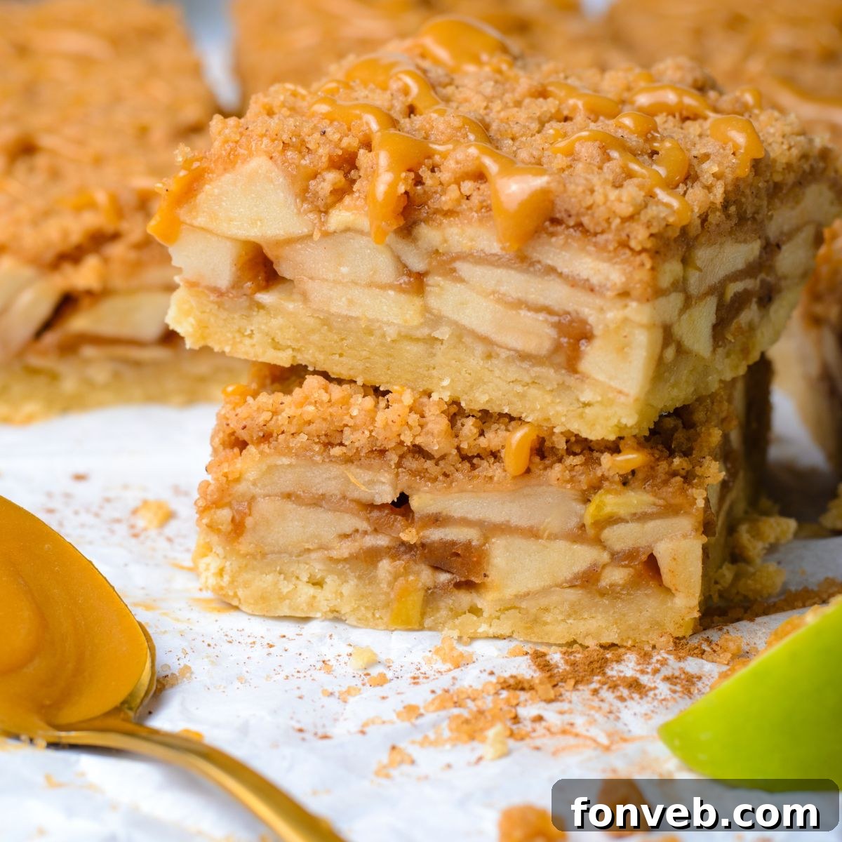 Caramel Apple Pie Bars stacked on top of one another with crumble around white table and a slice of green apple to side