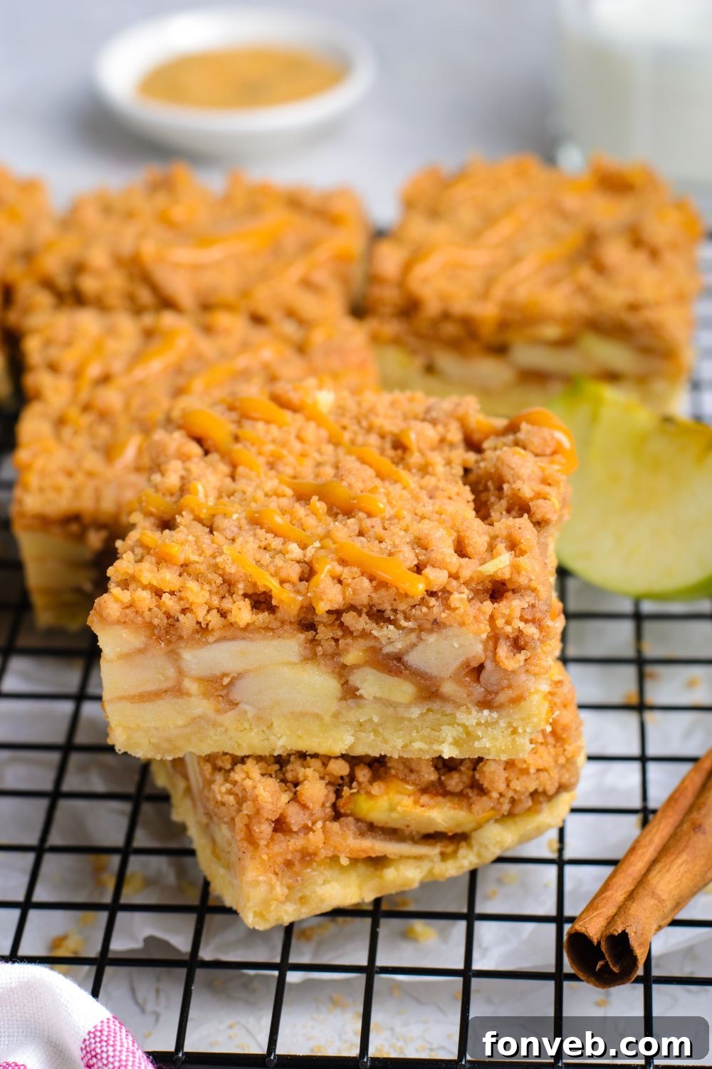 Caramel Apple Pie Bars stacked on top of one another and more bars around the cooling rack with cinnamon sticks and sliced apples