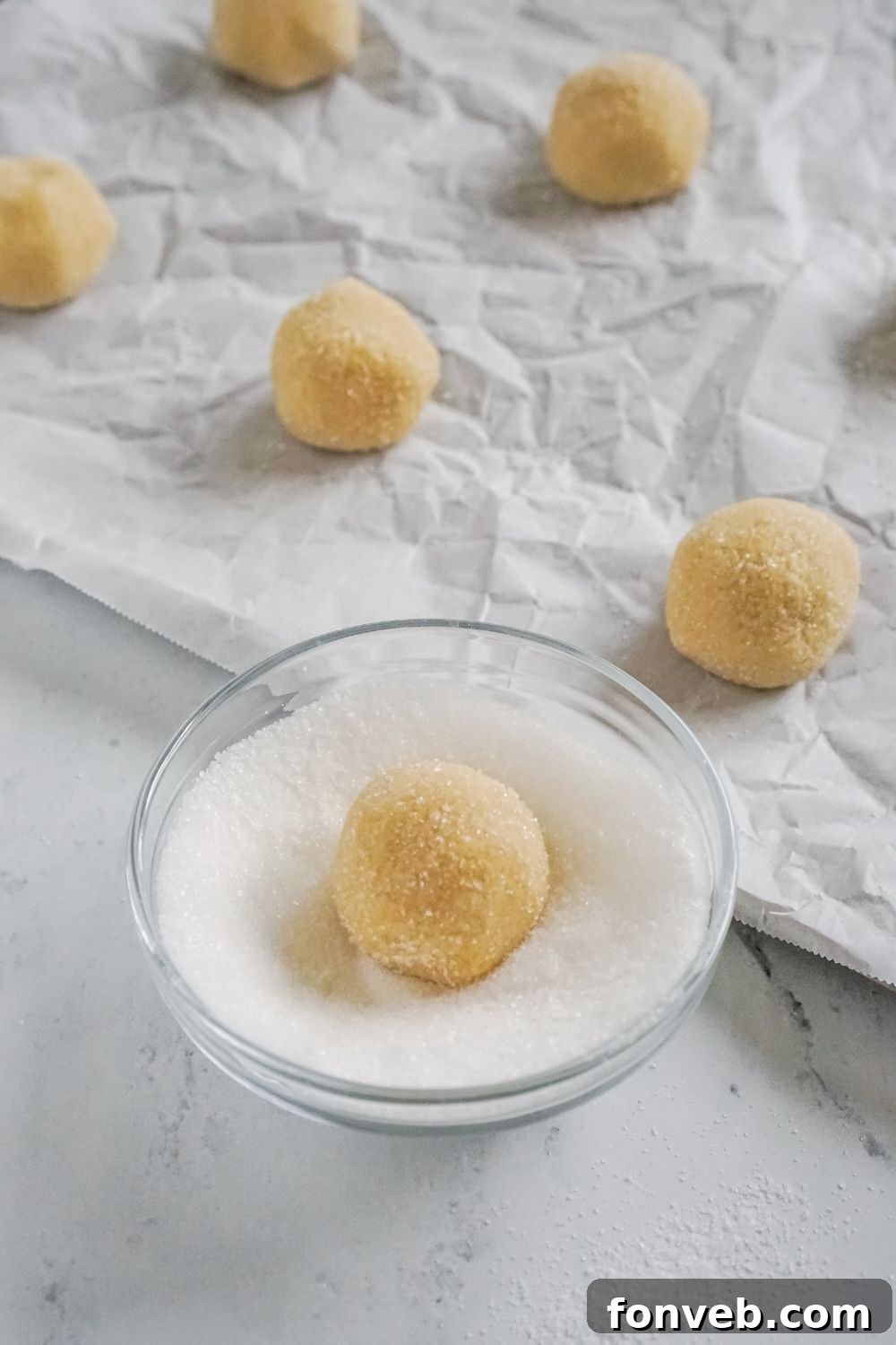cookie dough being rolled in a small bowl of sugar by baking sheet