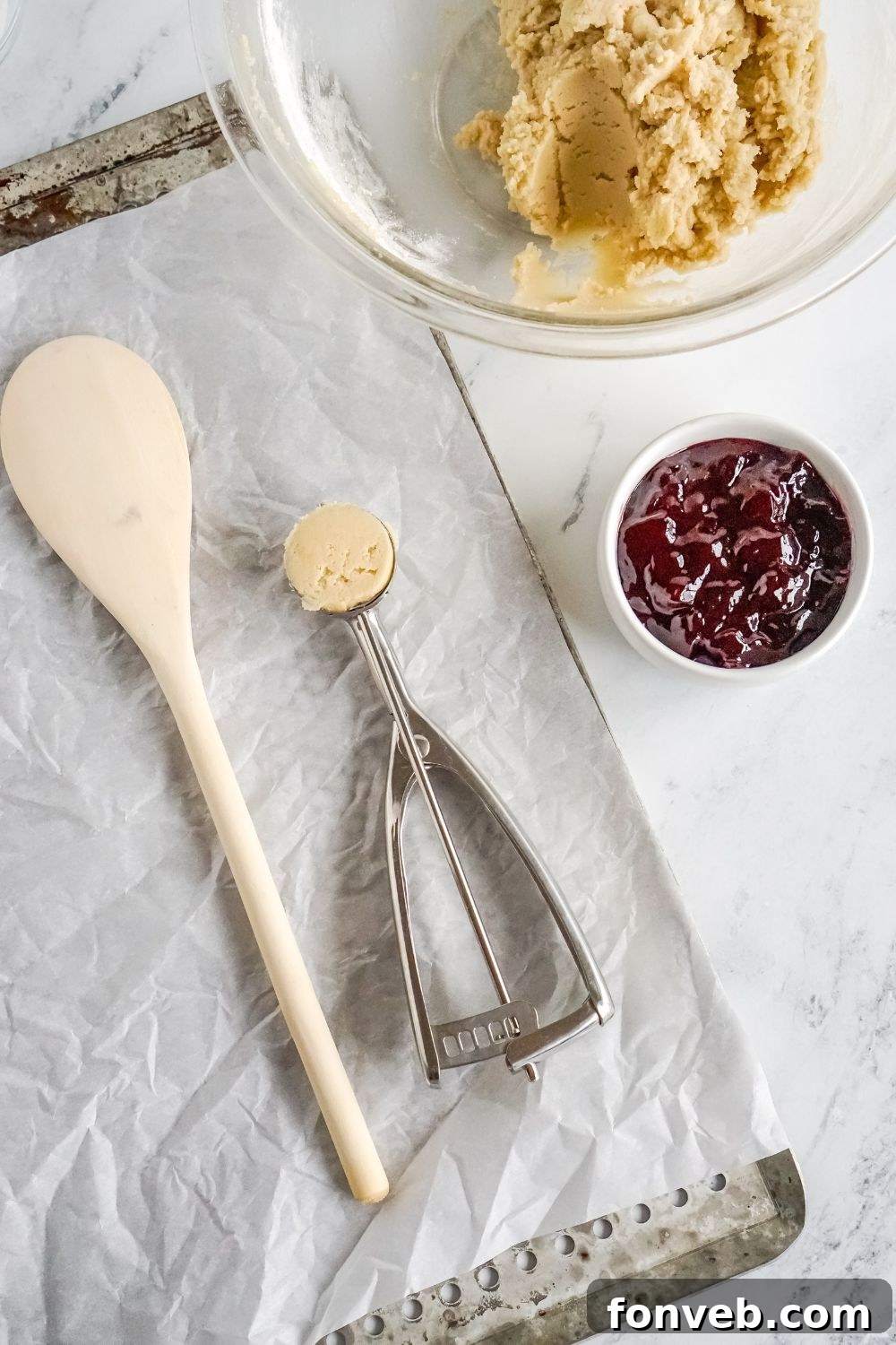 Raspberry Thumbprint Cookie dough in a glass bowl, with jam in a small bowl to side and cookie baking sheet on table