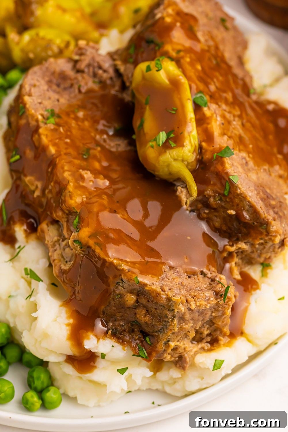 a plate with slices of meatloaf on it with a bed of mashed potatoes, peas, and gravy poured over the top of it
