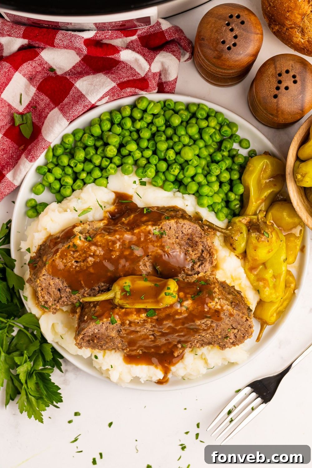 looking down on table to a plate full of Crockpot Mississippi Meatloaf with mashed potatoes, peas, and peppers to the side with a red checkered towel in the background 