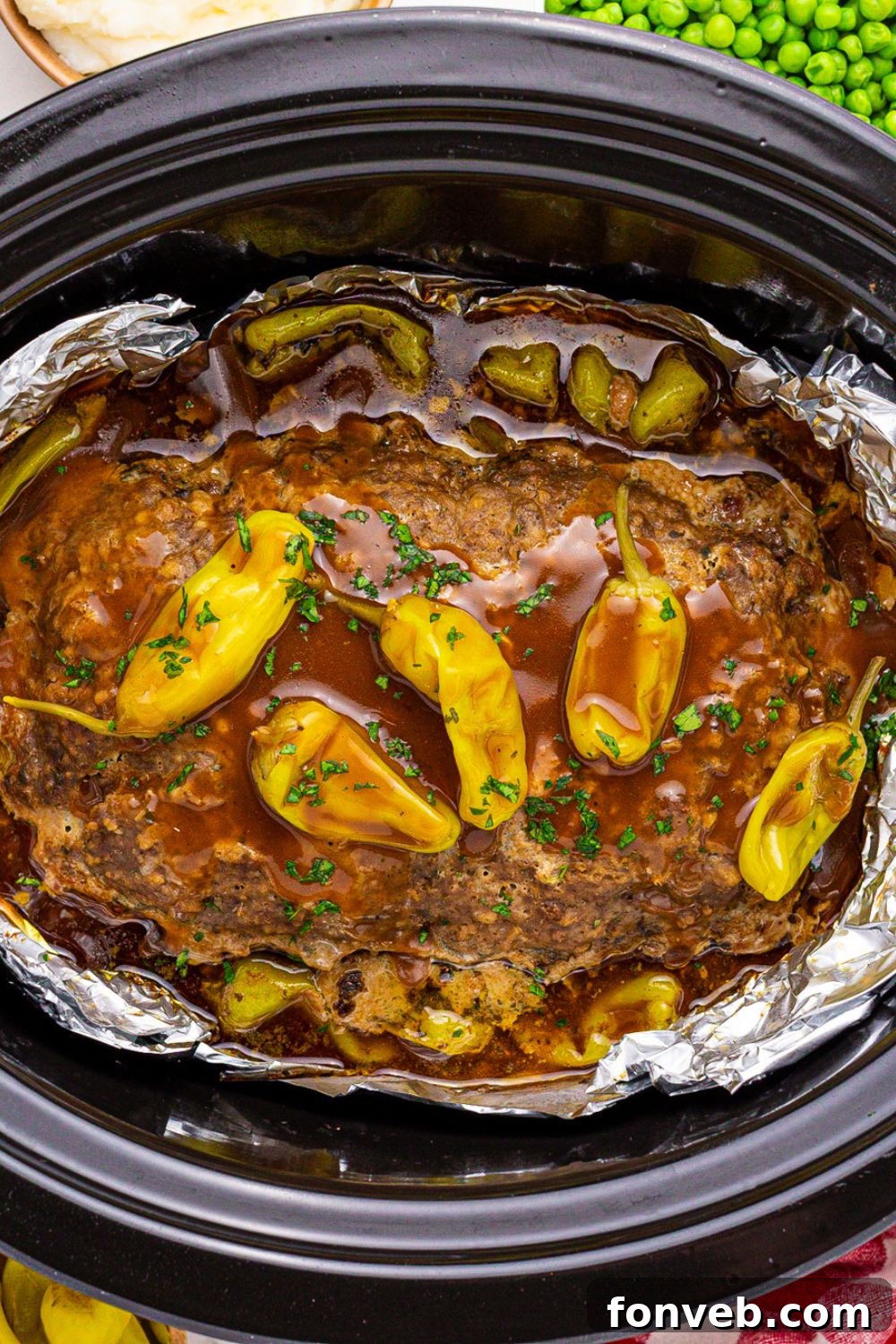 black slow cooker side ways on the table with meatloaf cooked in the slow cooker with gravy and peppers on top 