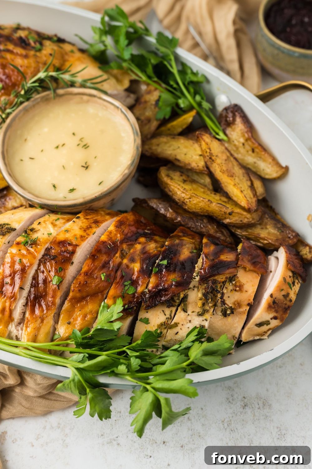 Side view of platter on white table with sliced roasted turkey on it, with a bowl of sauce and roasted potatoes