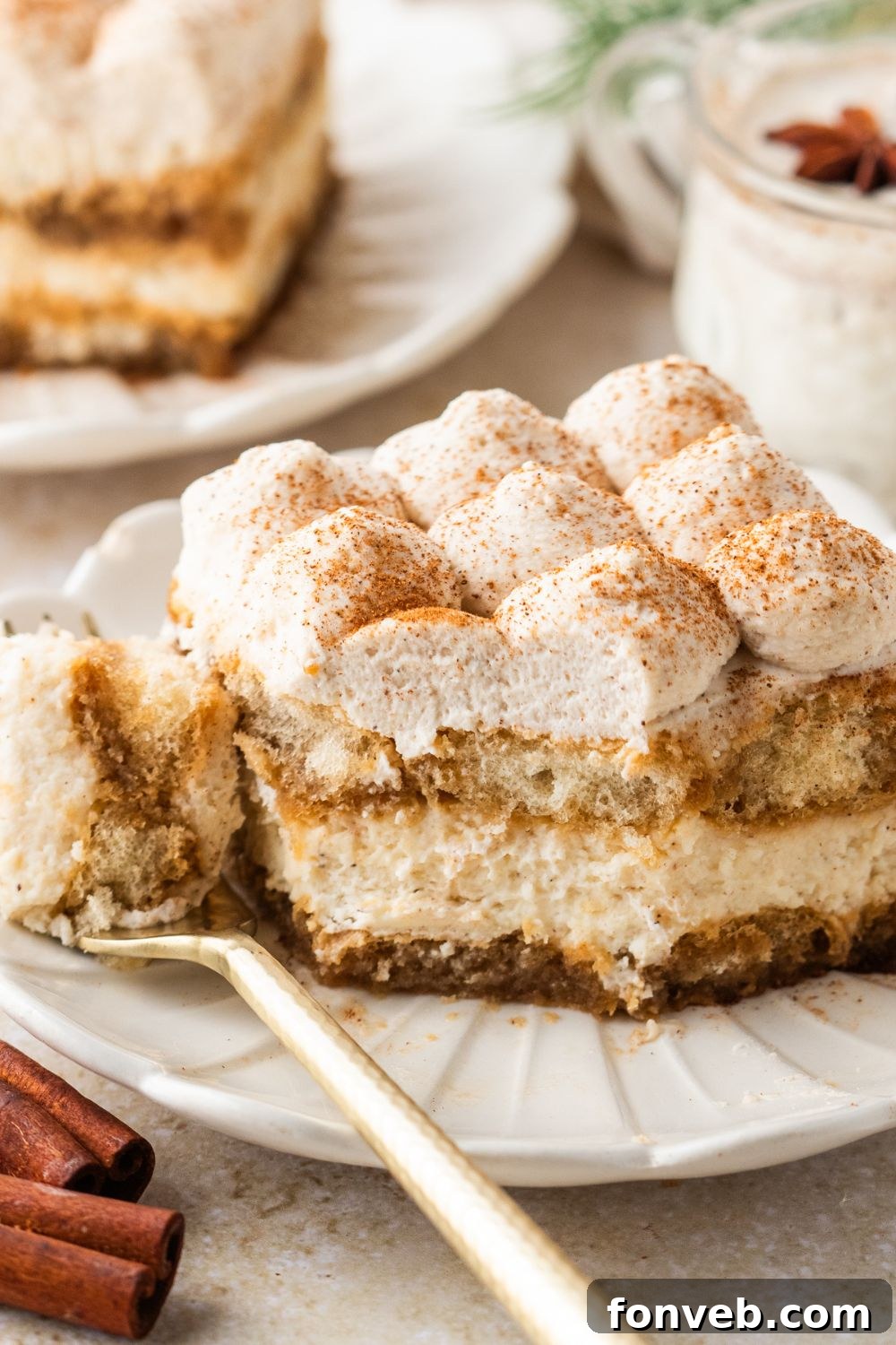 Eggnog Tiramisu sliced into a square on plate wit ha bite on a fork on side of plate with cinnamon sticks on table 