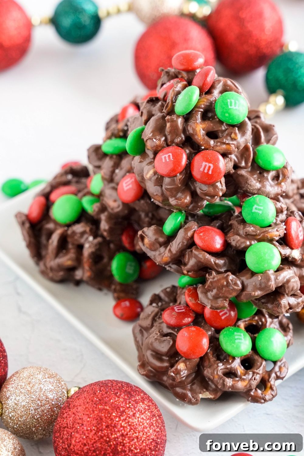 Sweet and Salty Slow Cooker Holiday Clusters 19 Crockpot Chocolate Pretzel Christmas Candy stacked on a white plate with Christmas ornament around the table