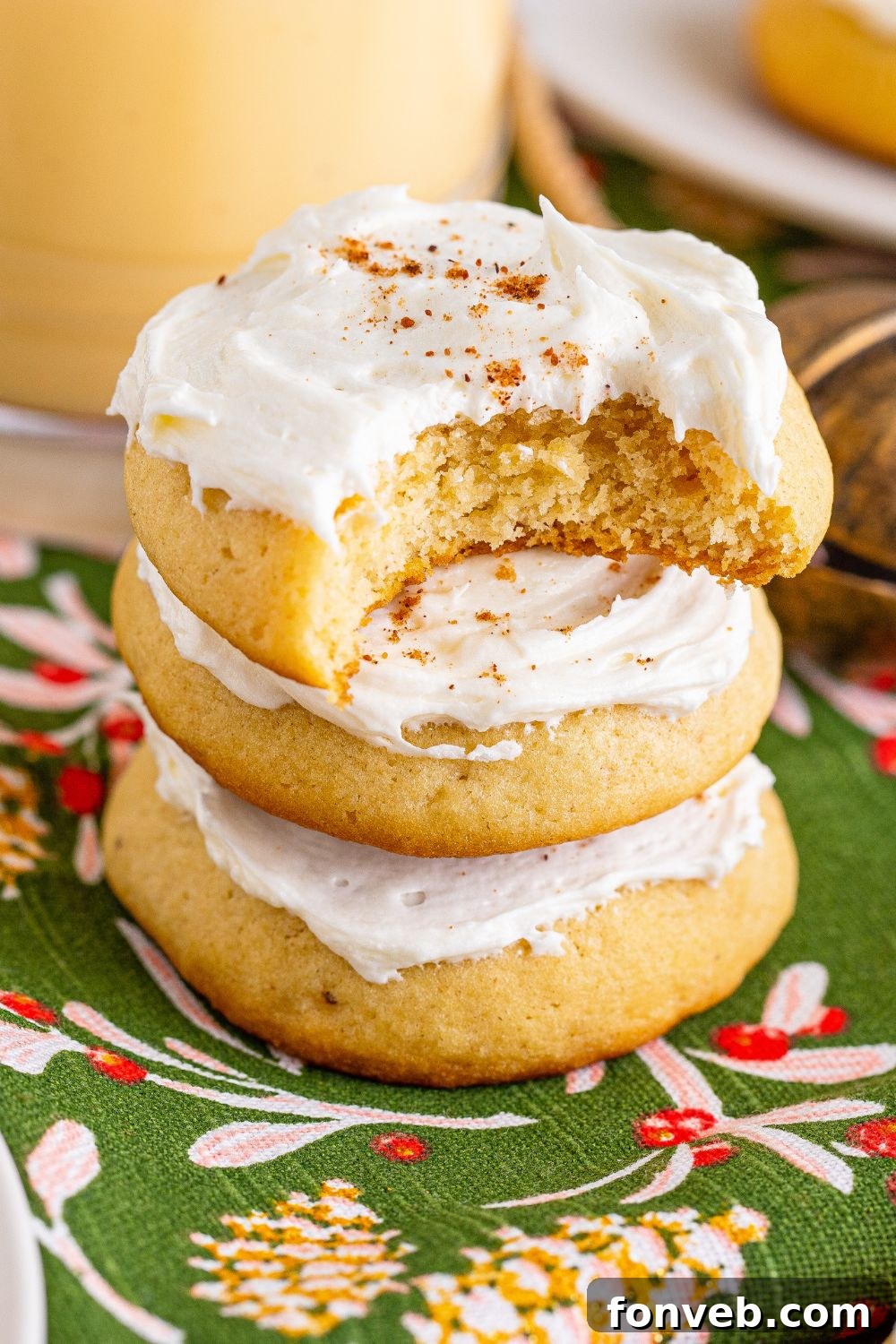 Soft Frosted Eggnog Cookies stacked on table with a bite taken out of it and a glass of eggnog to side of table