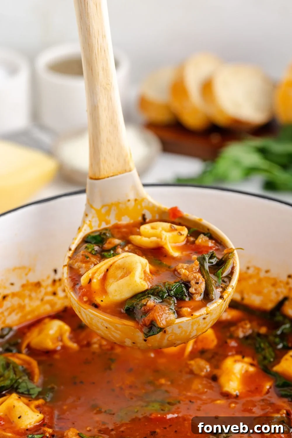A ladle expertly lifts a portion of Sausage Tortellini Soup from a pristine white pot on a table, highlighting the rich texture and ingredients