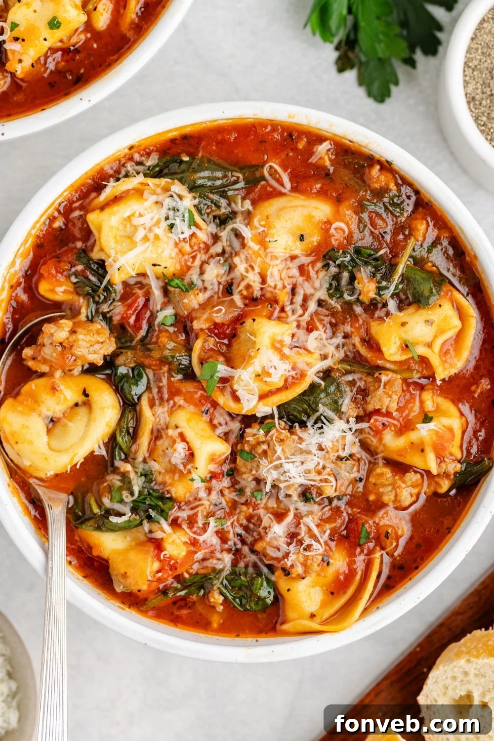 A pristine white bowl filled with enticing Sausage Tortellini Soup, accompanied by a spoon resting gracefully on the side, on a simple white table