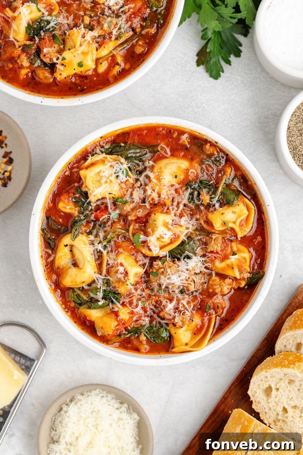 A beautifully set dinner table featuring multiple white bowls brimming with Sausage Tortellini Soup, generously topped with Parmesan cheese. A small bowl of extra grated cheese and a cutting board of sliced French bread complete the inviting spread.