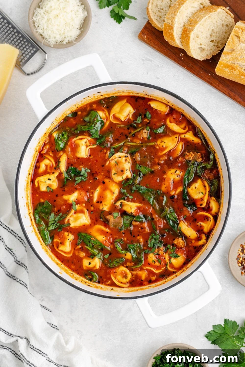 A rustic scene: a stockpot full of Sausage Tortellini Soup sits on a marble counter, accompanied by sliced bread and a bowl of grated Parmesan cheese, inviting a delicious meal
