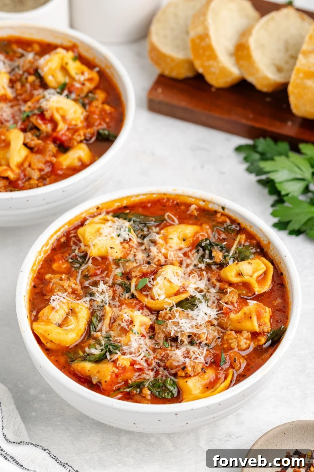 Multiple white bowls of Sausage Tortellini Soup are elegantly arranged on a table, accompanied by a cutting board laden with freshly sliced French bread, inviting a cozy dinner