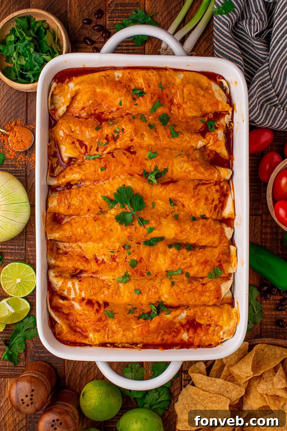 white 9x13 casserole dish on wooden table full of Steak Enchiladas with taco toppings around the pan 
