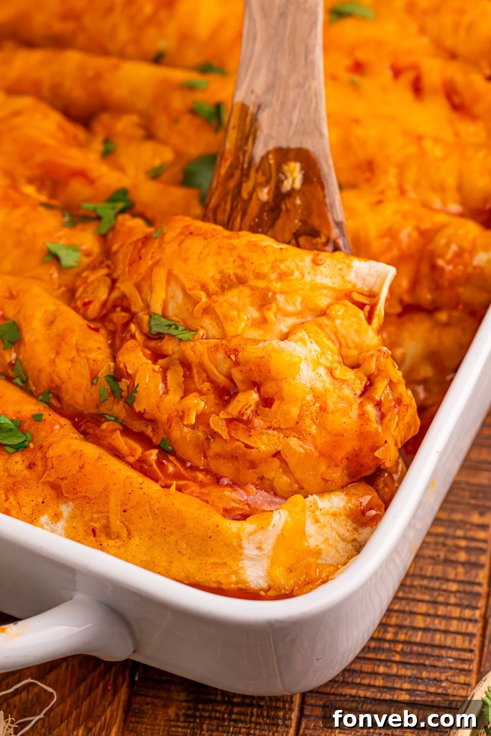 Steak Enchiladas in a white casserole dish with a wooden spoon scooping up an enchilada our of the pan 