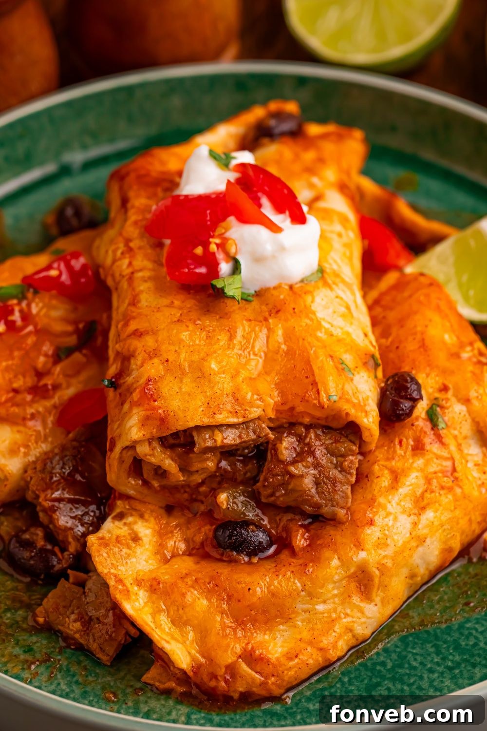 Steak Enchiladas on a plate with some sour cream and salsa on top with a few limes to side of the plate on wooden table 