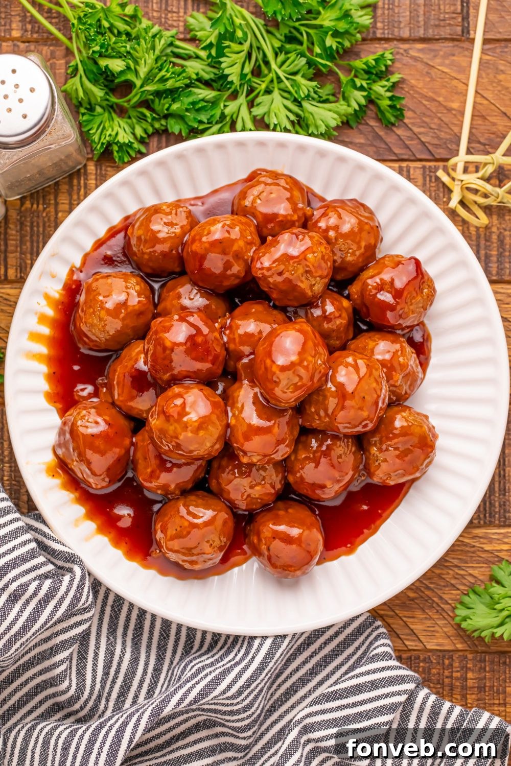 a wooden table with a white plate full of peach bbq meatballs, and salt and pepper to side, parsley, and toothpicks for serving up 