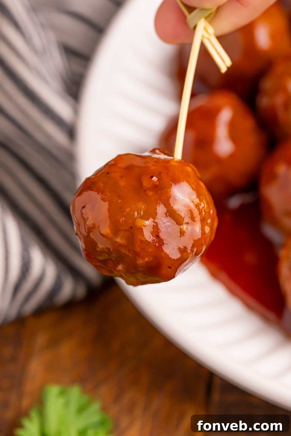 toothpick being held with a Peach BBQ Meatball on it and a plate full of more meatballs below on table 