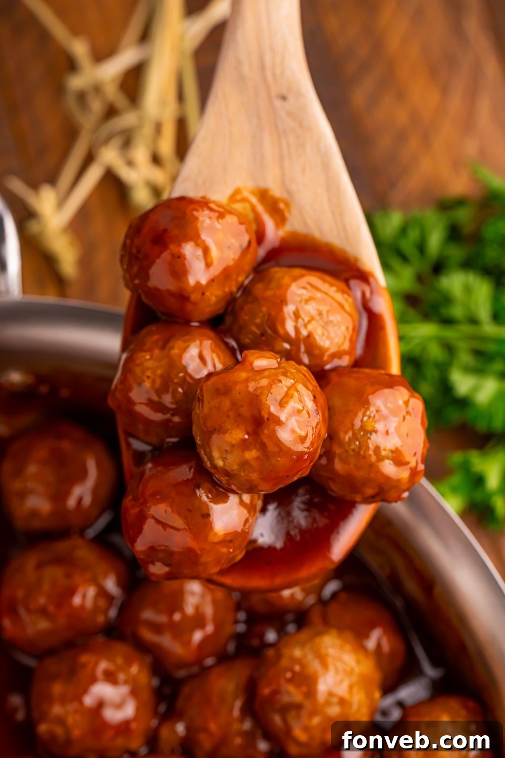 Peach BBQ Meatballs in a skillet with a wooden spoon full of meatballs and a bunch of parsley to side of skillet on wooden table 