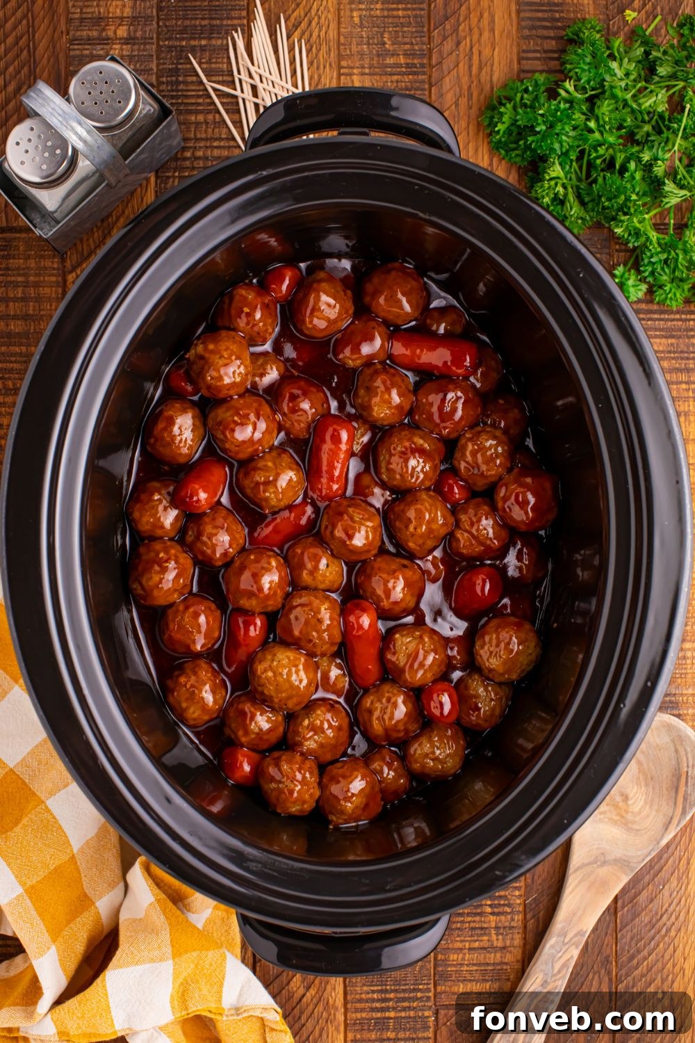 looking down on table to see a black crockpot full of Crockpot Teriyaki BBQ Cocktail Meatballs & Lil Smokies with salt and pepper shaker to side and a wooden spoon in front 