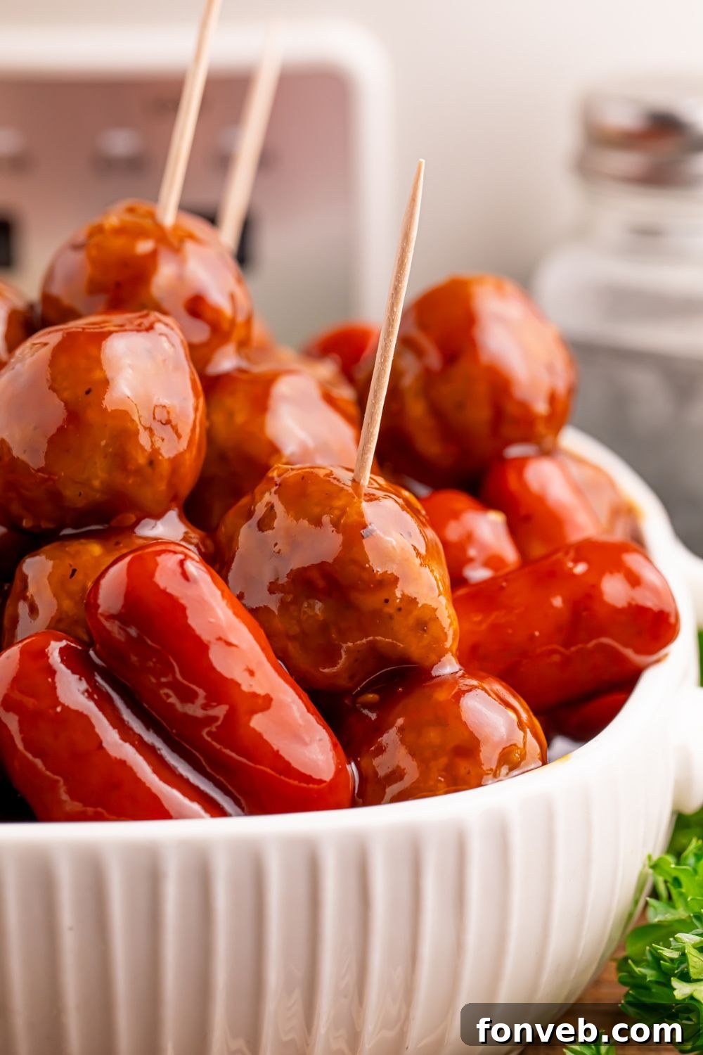 Crockpot Teriyaki BBQ Cocktail Meatballs & Lil Smokies in a white bowl on table with toothpicks poked into a few of them with the silver crockpot behind it on the table 
