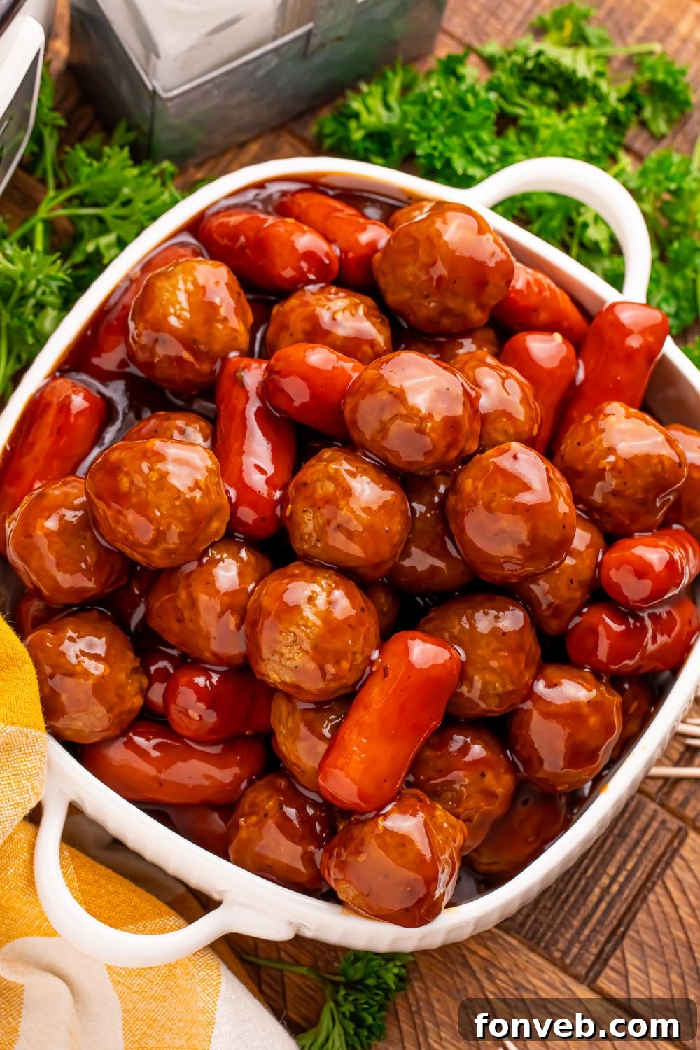 looking down on table to see a container full of Crockpot Teriyaki BBQ Cocktail Meatballs & Lil Smokies with some fresh parsley around the table and a yellow towel to the side 