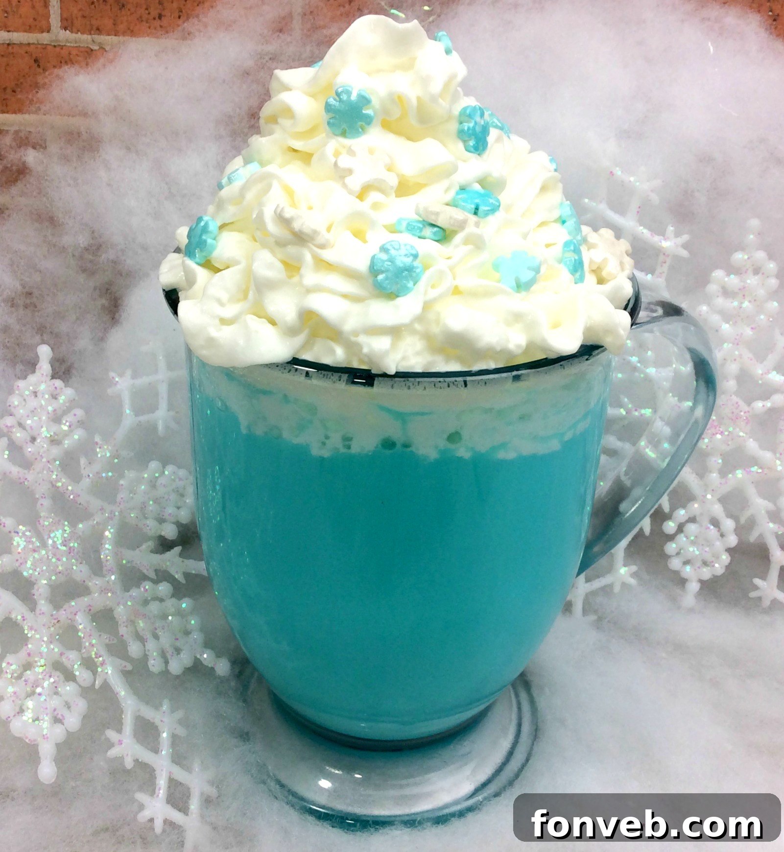 A steaming mug of vibrant blue hot cocoa, topped with fluffy white whipped cream and festive blue and silver sprinkles. The mug is held by hands and set against a soft, winter-themed background.
