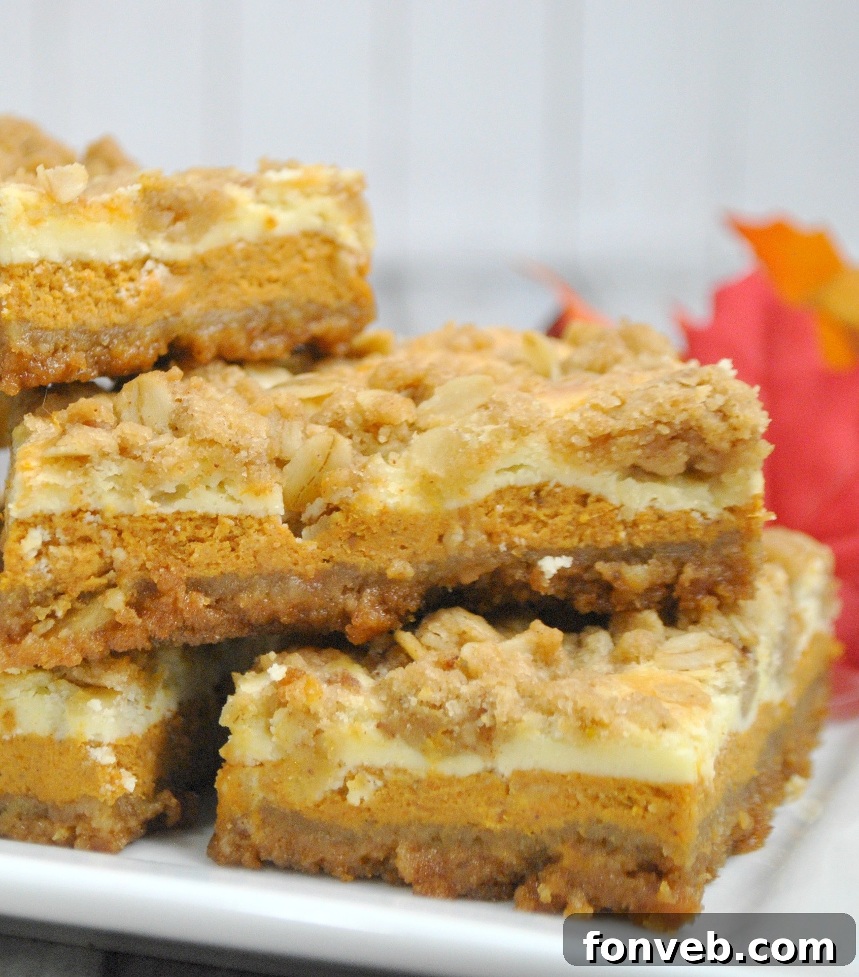 Harvest Pumpkin Crumble Cheesecake Squares 6 A charming stack of three pumpkin cheesecake bars on a white plate, subtly garnished with cinnamon, ready to be served and enjoyed.