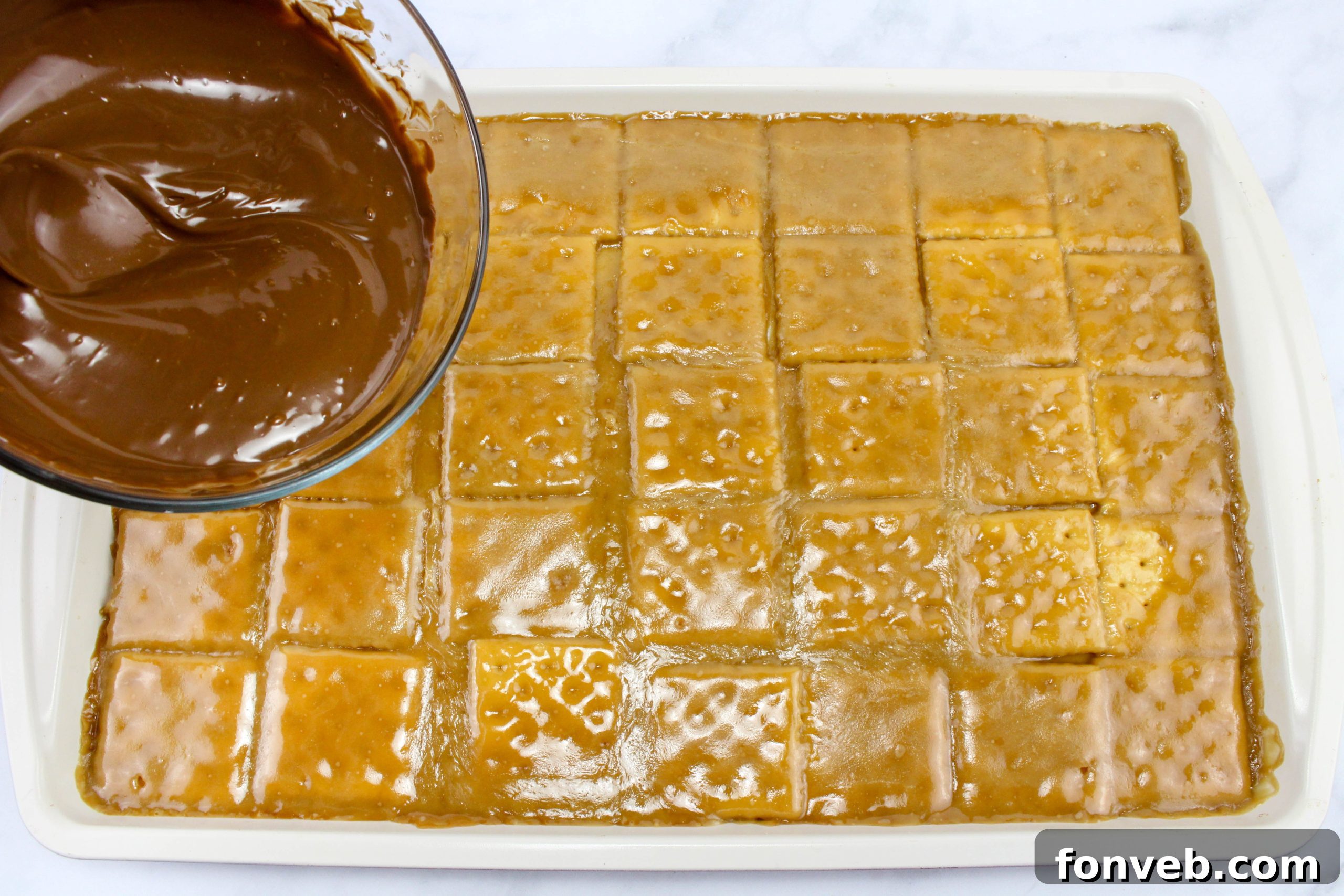 Melted milk chocolate chips being poured and spread evenly over the caramel-coated saltine crackers on a baking sheet.