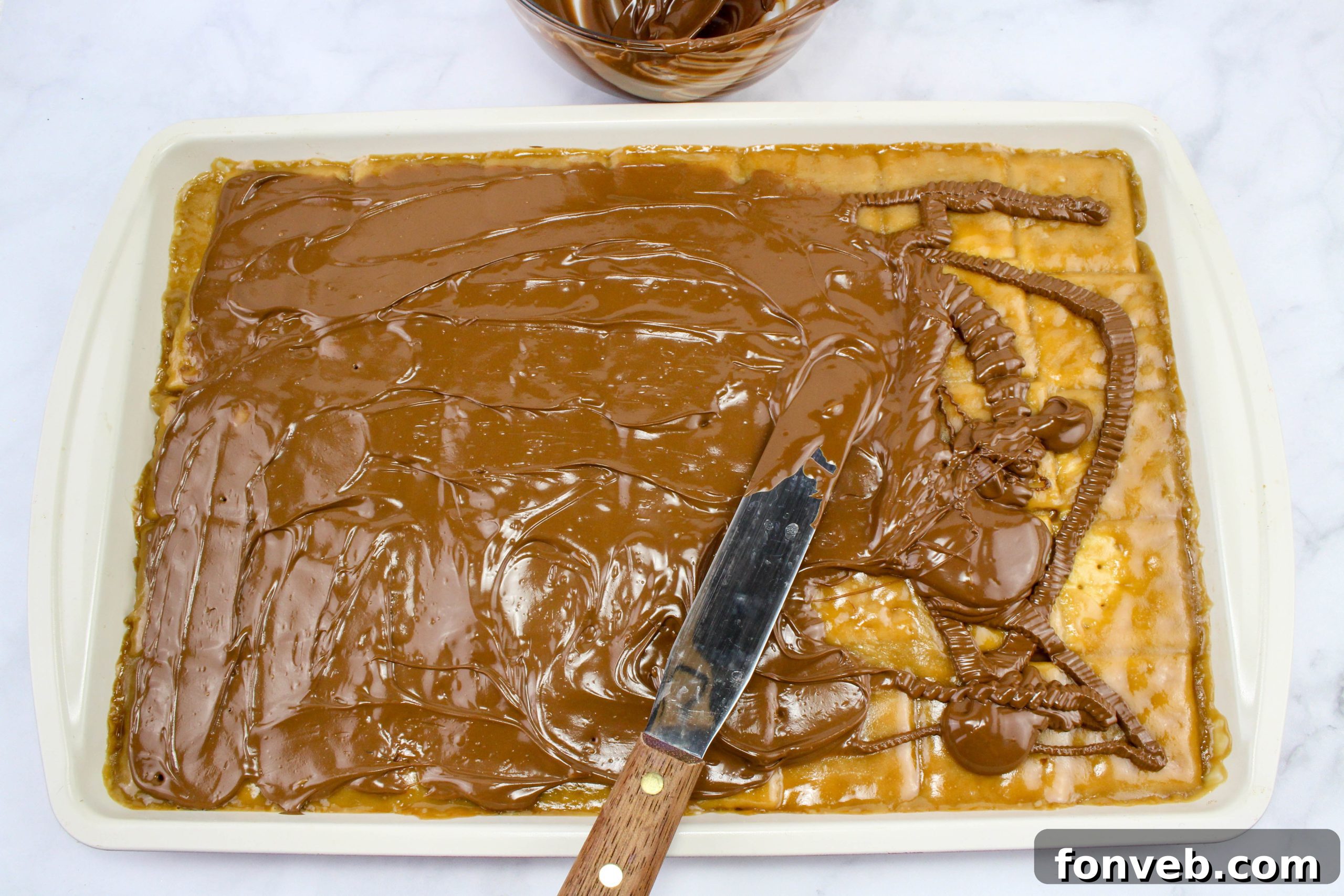 A spatula smoothing the melted chocolate layer over the caramel crackers, preparing for the toppings.