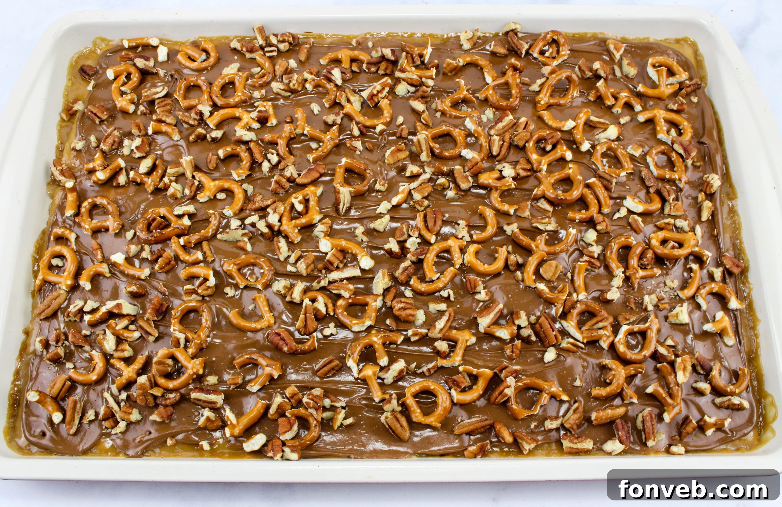 Chopped pretzels being sprinkled generously over the still-warm melted chocolate layer on the cracker toffee.