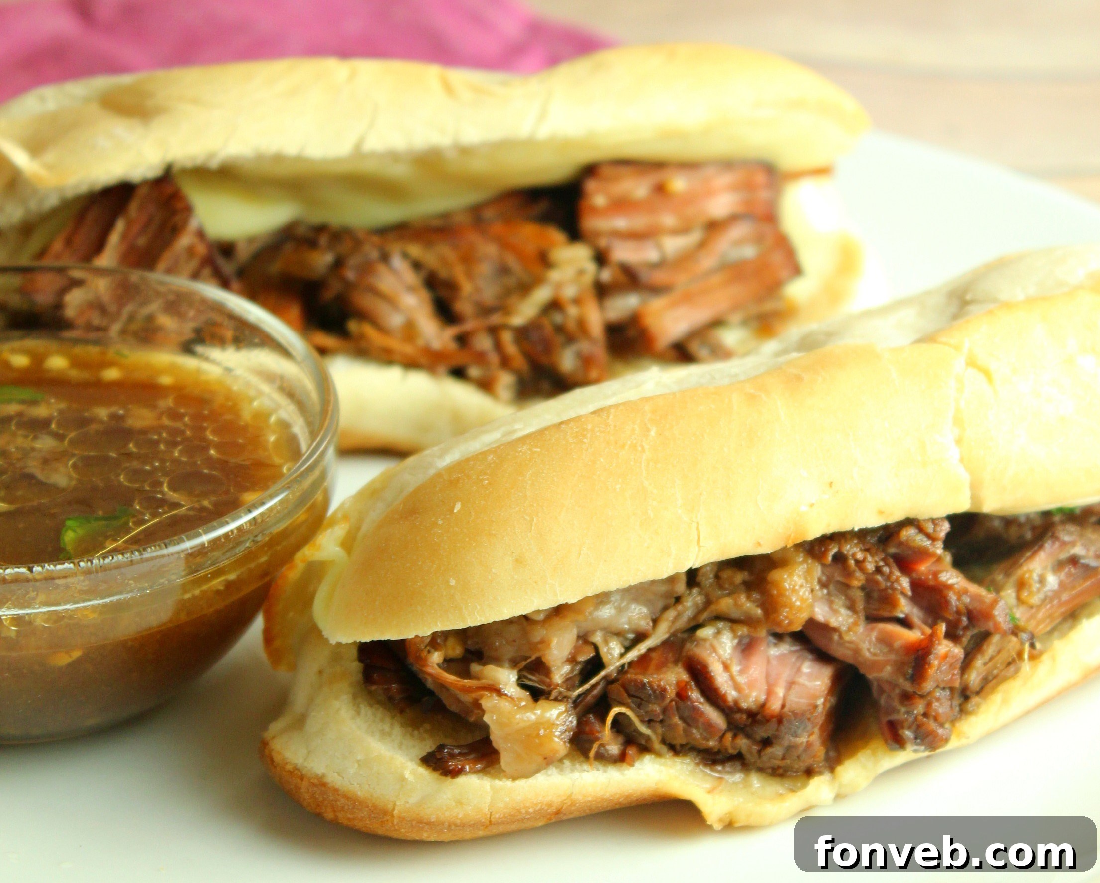 Two French Dip sandwiches, one with a close-up on the melted cheese, next to a bowl of rich au jus for dipping.