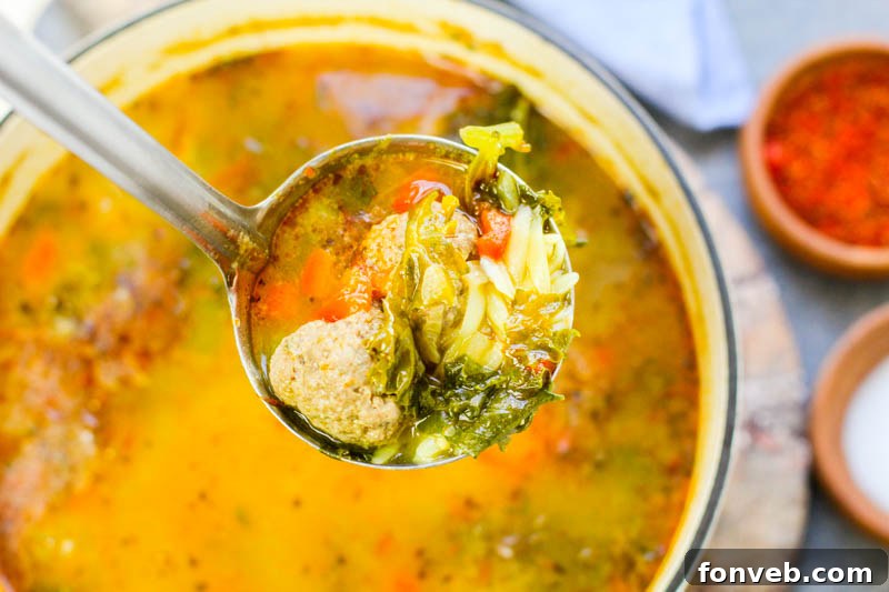 Rustic Tuscan Meatball Soup 6 A rustic wooden spoon resting in a large bowl of Tuscan Meatball Soup, filled with savory broth, meatballs, pasta, and fresh kale, on a light-colored background.