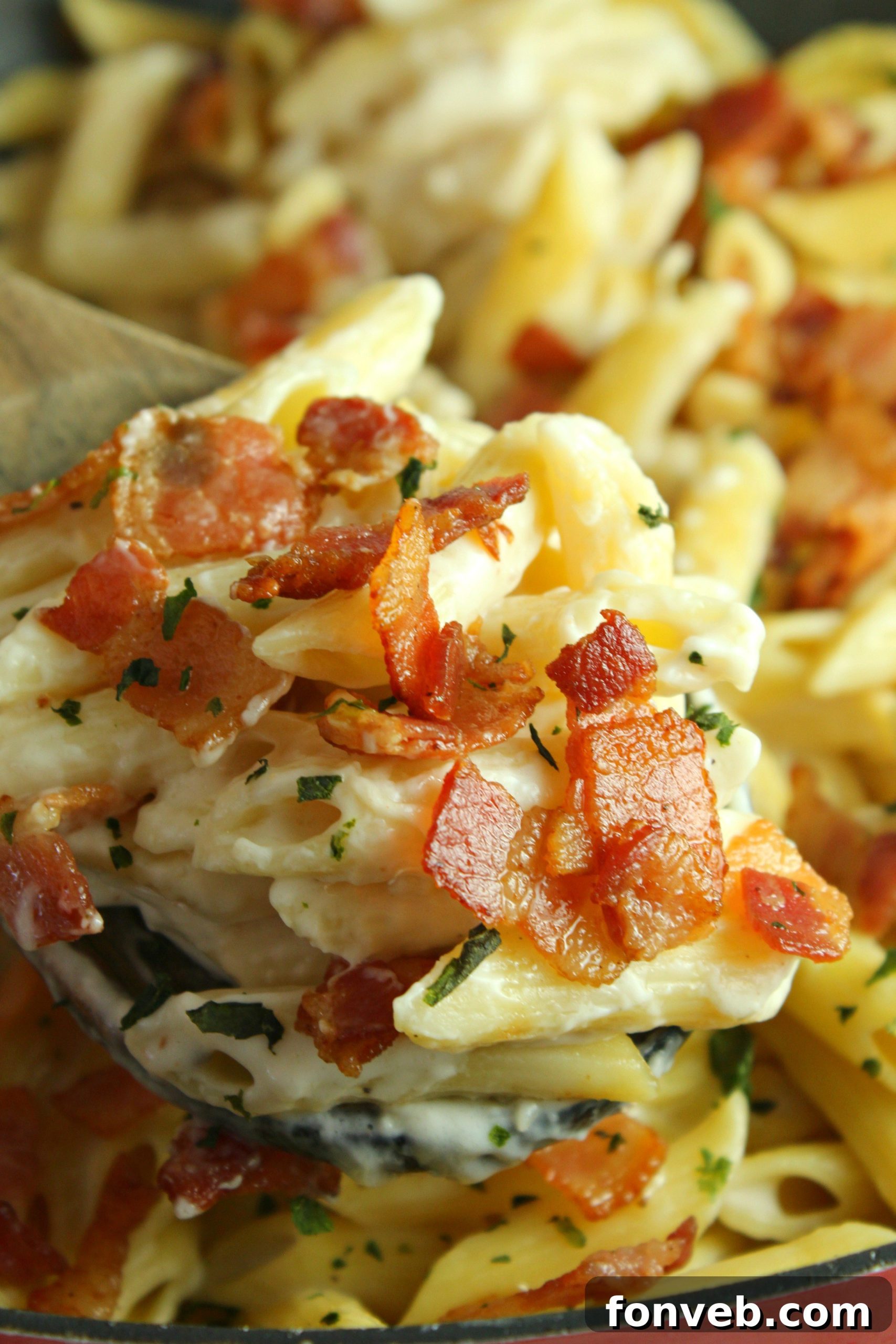 A hand holding a fork lifting a mouthful of creamy bacon alfredo pasta from a bowl, steam gently rising.
