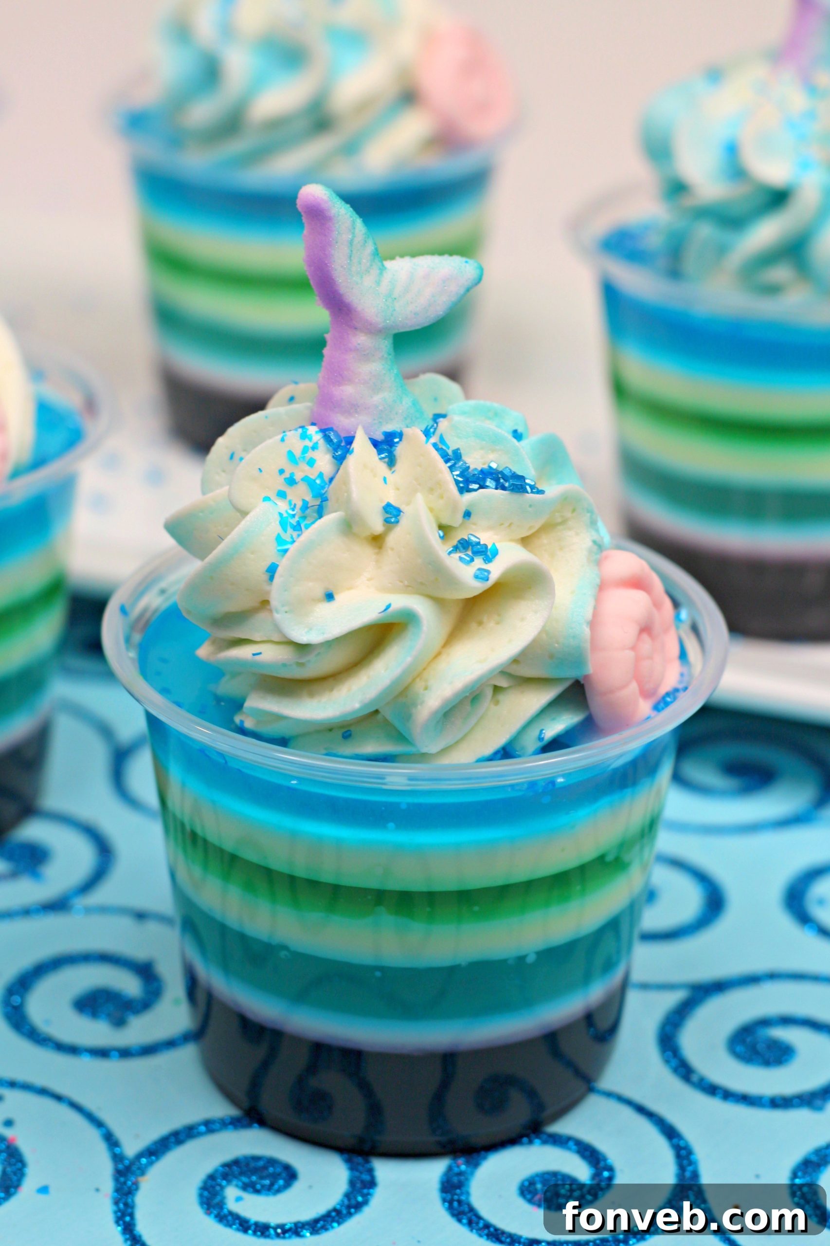 Close-up of vibrant Mermaid Jello Shots stacked in shot glasses, showing purple, blue, white, and green layers, topped with whipped cream and mermaid-themed decorations.