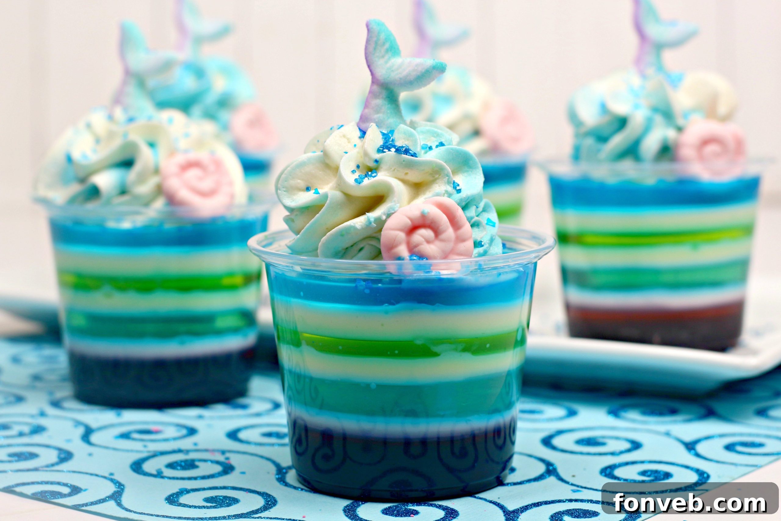 A close-up of a single Mermaid Jello Shot, perfectly layered with purple, white, blue, and green, topped with whipped cream and a small mermaid tail decoration.
