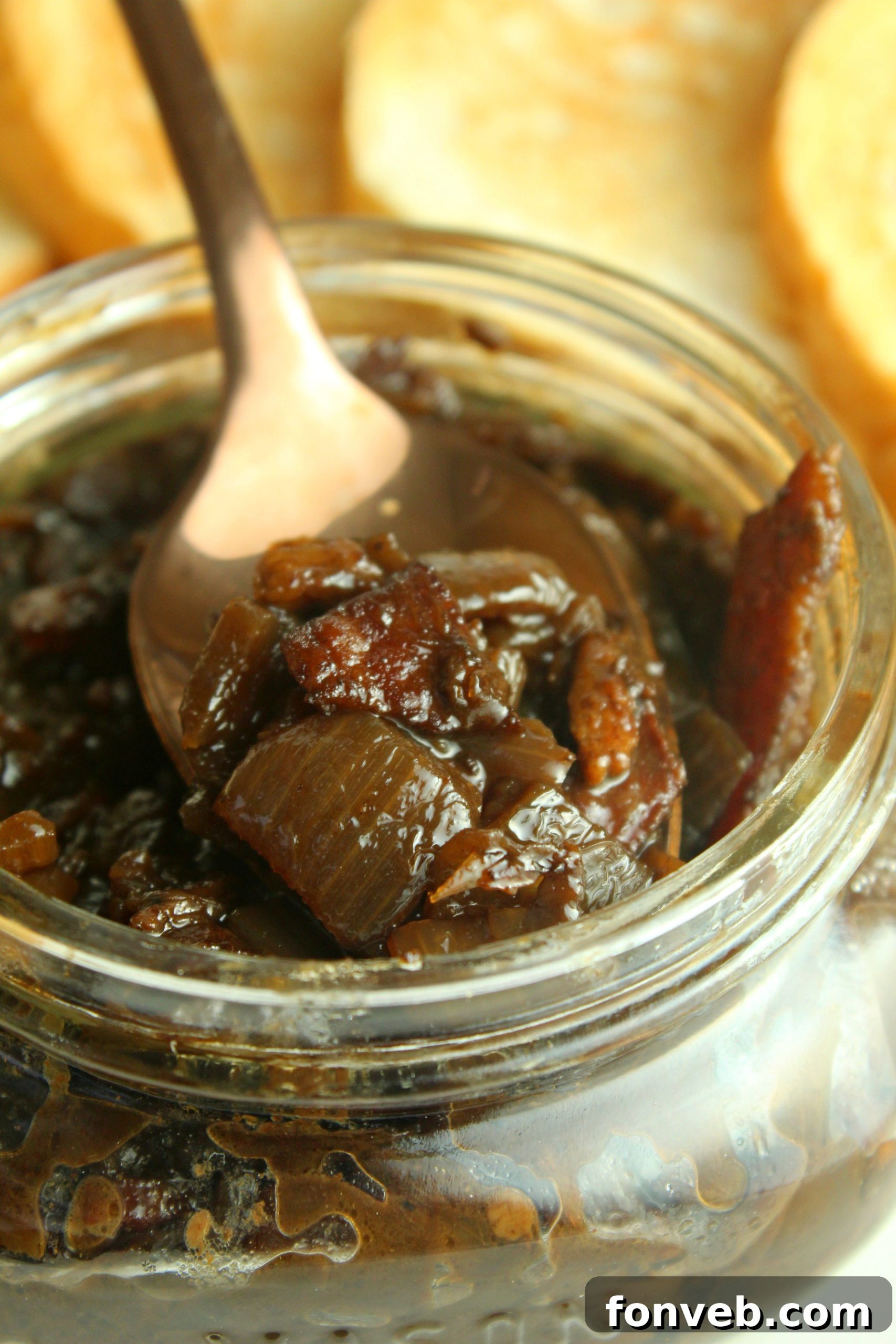 A close-up of a spoon lifting a sticky, glossy portion of bacon jam from a bowl, highlighting its rich texture.