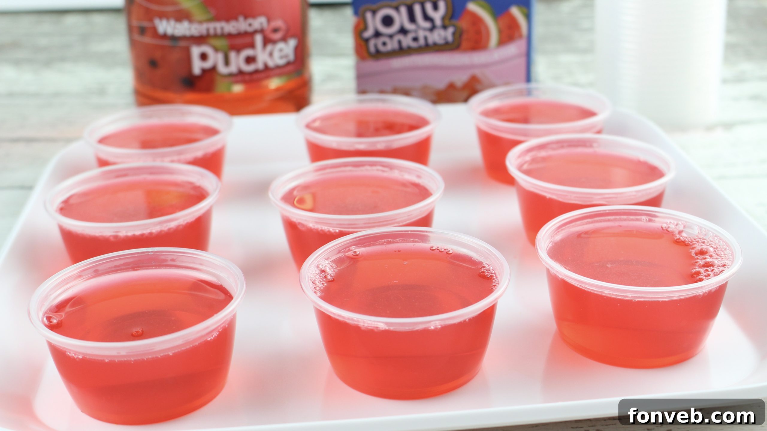 A hand holding a clear plastic cup filled with bright green Jolly Rancher Jello, with other colorful shots in the background.
