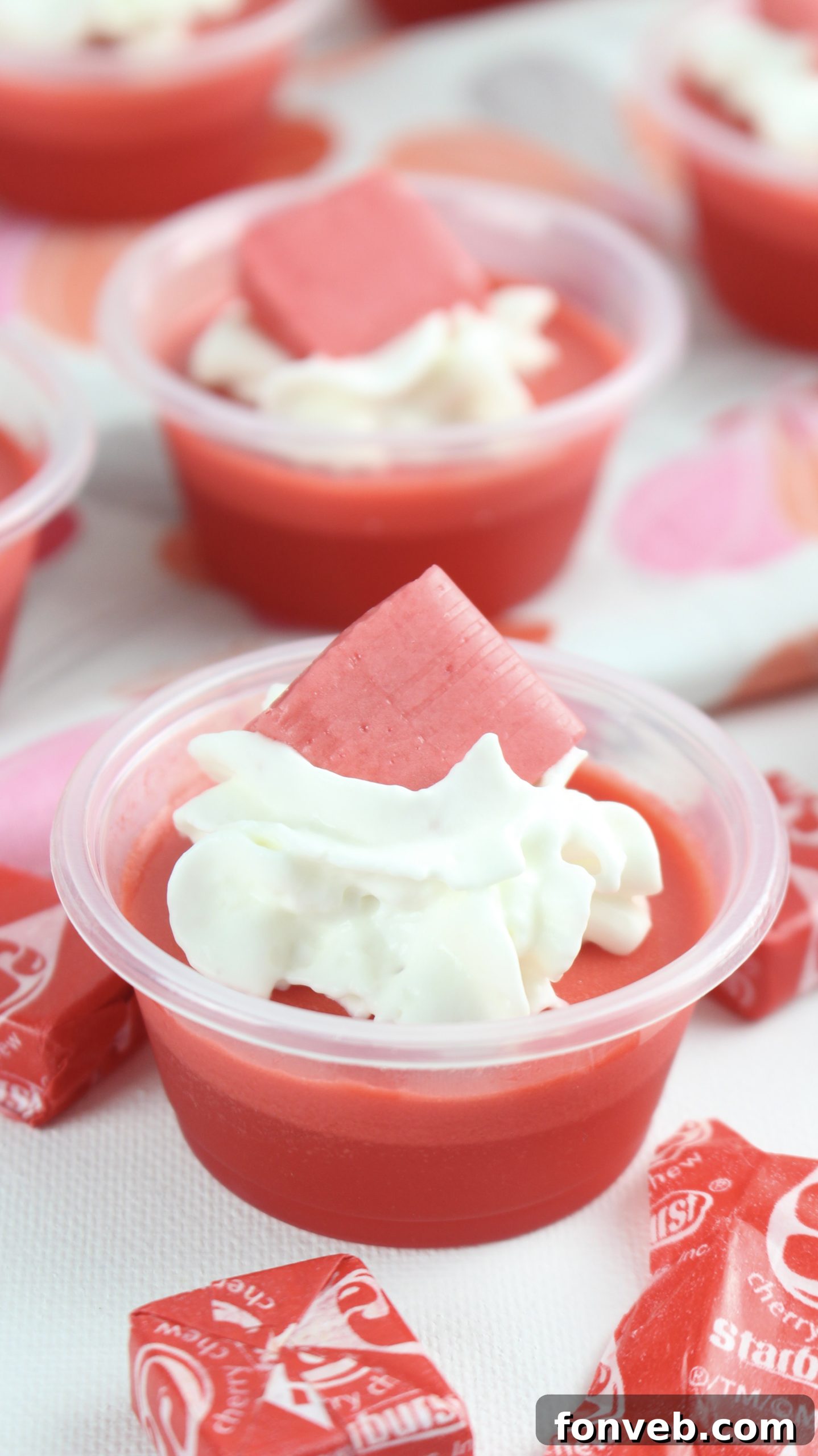Close-up of vibrant Cherry Starburst Jell-O Shots in small plastic cups, garnished with whipped cream and a cherry Starburst candy.