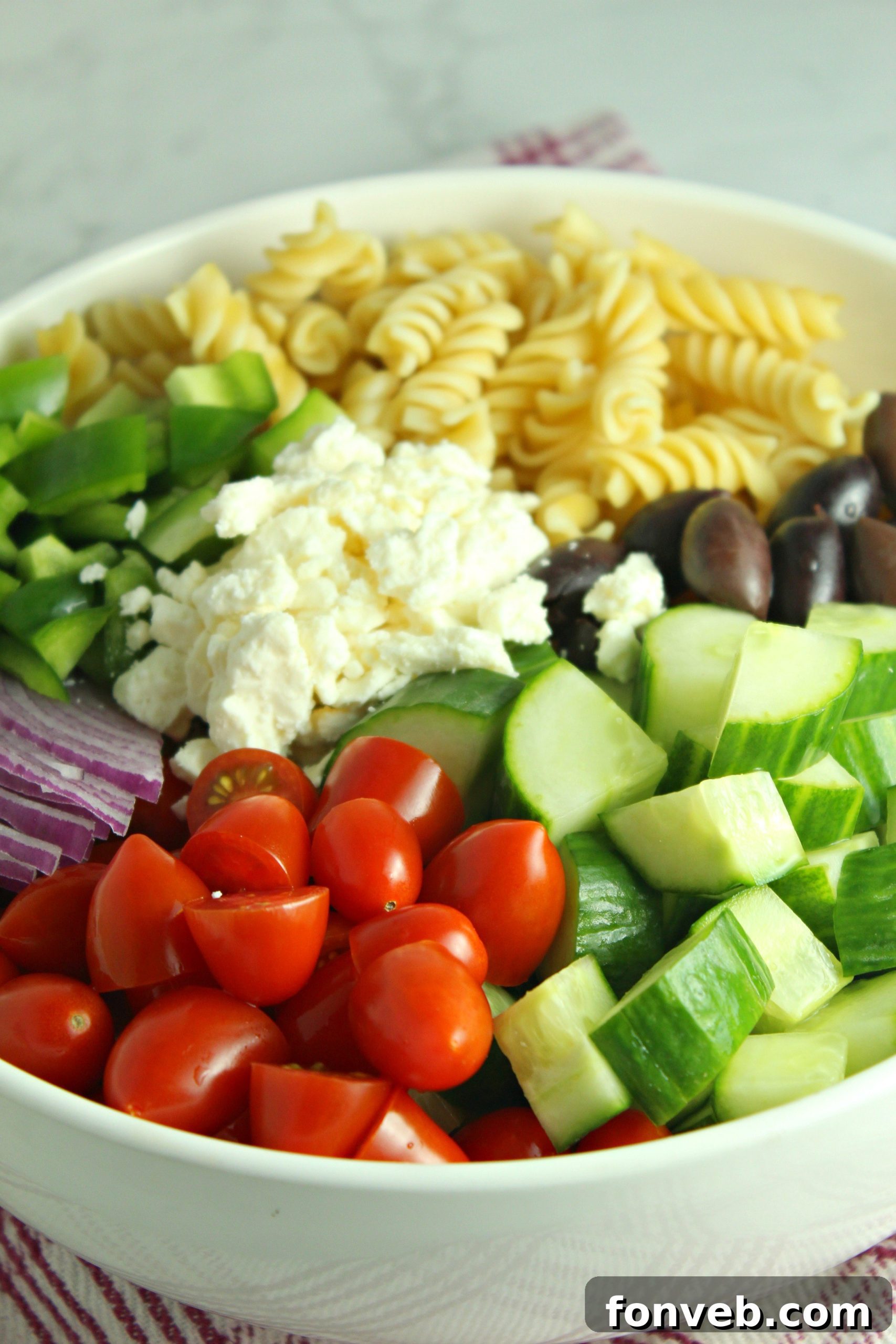 A flat lay image of Greek Pasta Salad ingredients, including pasta, cucumber, tomatoes, olives, feta, and parsley, ready for preparation.