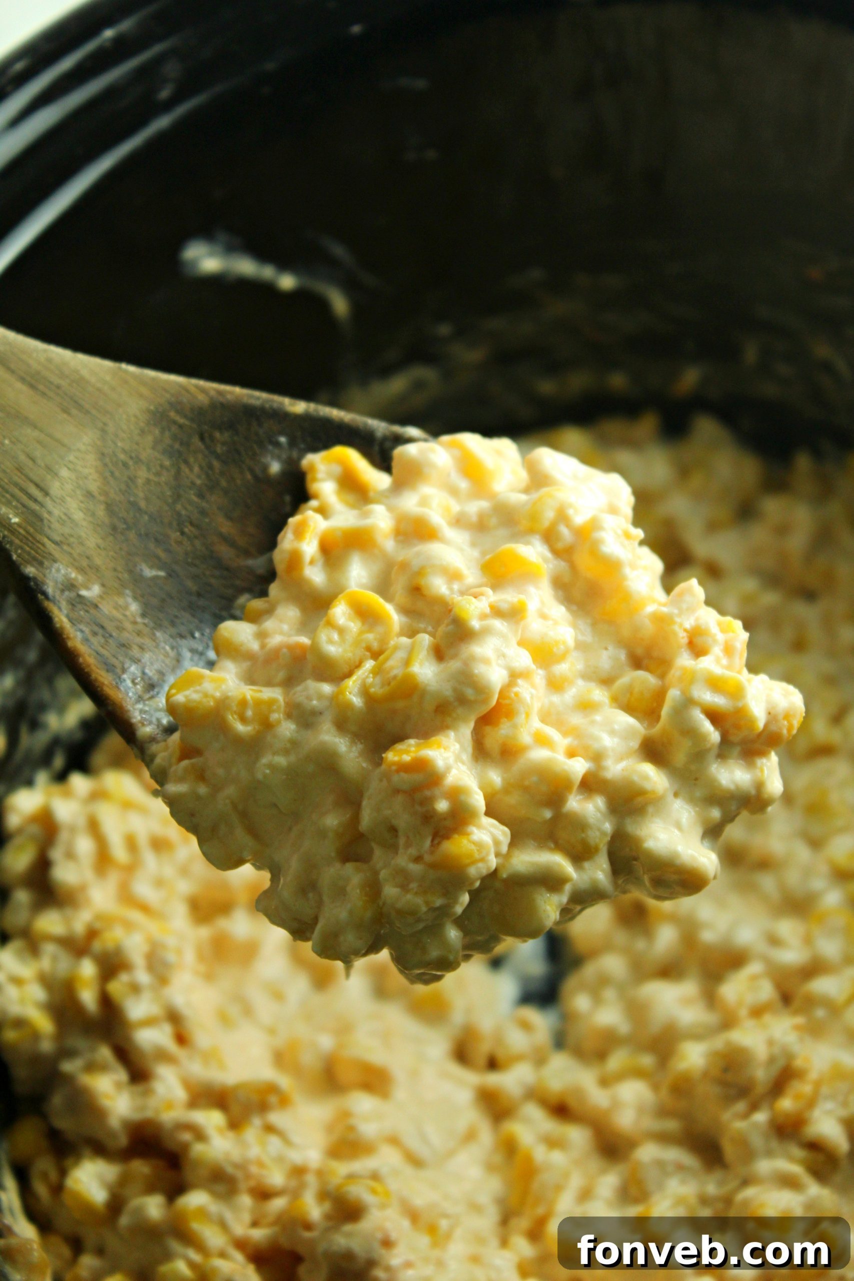 Serving spoon scooping creamy slow cooker cheesy corn from a large bowl, emphasizing its creaminess.